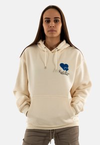 Sudadera con capucha color crema, con cordones, bolsillo frontal y un logo de corazón azul con texto en la parte izquierda del pecho. Textura de tela suave.