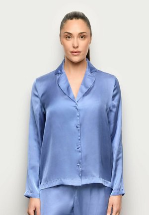 Mujer con cabello oscuro recogido en una coleta lisa, vestida con un conjunto de pijama azul claro de satén de manga larga y abotonado, sobre un fondo claro y liso.