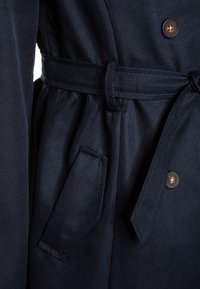 Námořnický modrý trenčkot s páskem, zapínáním na knoflíky a boční kapsou. Materiál se zdá být hladký s mírným leskem.