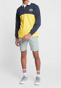Camisa polo de manga larga con parte superior azul oscura y parte inferior amarilla, cuello blanco, con un logo. Combinada con pantalones cortos verdes claros y calcetines a rayas.
