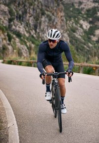 Un cycliste portant un maillot ajusté bleu marine à manches longues et un short noir roule sur un vélo de route noir élégant sur une route asphaltée sinueuse, avec un casque blanc.