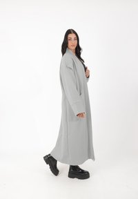 Langer, hellgrauer Mantel mit weiten Ärmeln, seitlichen Taschen und geradem Schnitt, kombiniert mit schwarzen klobigen Stiefeln. Glatte Textur.