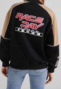 Osoba oblečená v černé a béžové bundě s nápisem "RACE DAY VIBES" a grafikou šachovnicové vlajky na zadní části a rukávu, doplněná modrými džíny.