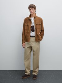 Brown suede jacket with two front pockets, white t-shirt beneath, beige wide-leg trousers, and dark brown suede shoes with rubber soles.