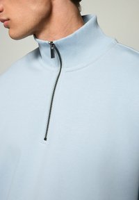 Lyseblå halv-zip trøje med ribbet krave og glat tekstur. Har en kontrasterende sort lynlås og minimalistiske designdetaljer.