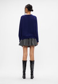 Tengerészkék, oversized kapucnis pulóver, pliszírozott szürke szoknya és térd felett érő fekete magassarkú csizma. Sima anyag, kerek dekoltázs és bordázott mandzsetta.