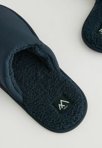 Pantoufles bleu marine avec une doublure douce et texturée et un matériau extérieur durable. Présentent un dos fermé et une étiquette de logo sur la semelle intérieure.
