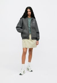 Γκρι φούτερ με φερμουάρ και μπροστινές τσέπες πάνω από πράσινη κορυφή, συνδυασμένο με μπεζ φούστα. Λευκά αθλητικά παπούτσια με πράσινες λεπτομέρειες. Λείος υφάσματος.