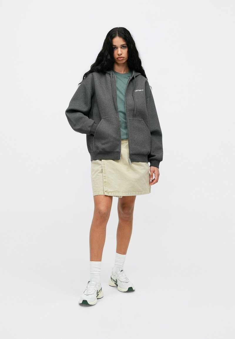 Grå zip-up sweatshirt med forlommer over en grøn top, parret med en beige nederdel. Hvide sneakers med grønne detaljer. Glat tekstur.