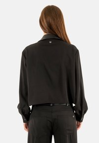 Mujer con cabello largo castaño, vestida con un top negro recortado y pantalones negros, de pie de espaldas frente a un fondo claro y liso.