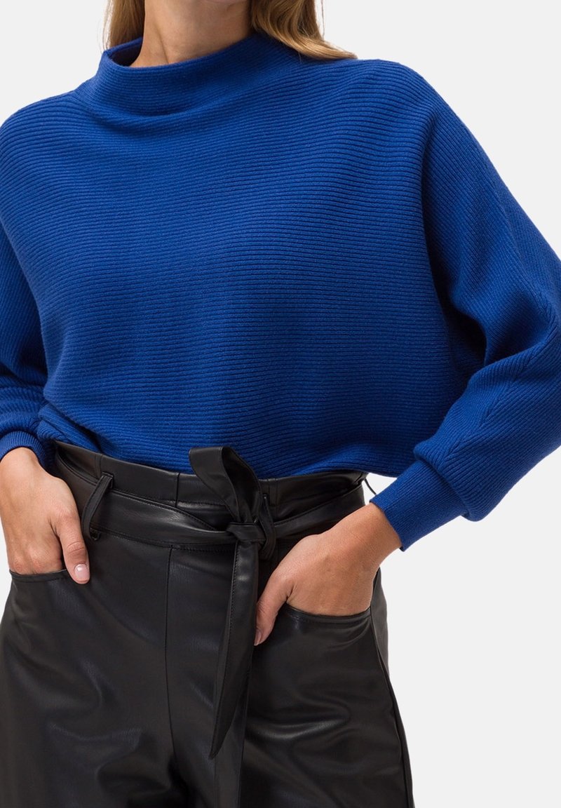 Femme portant un pull côtelé bleu rentré dans un pantalon en cuir noir taille haute avec une ceinture nouée, mains dans les poches, sur fond blanc.