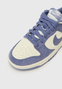 Nike tenisice svijetloplavog nubuka s kremastim kožnim prednjim dijelom. Značajke uključuju ljubičaste vezice i teksturiranu gumenu potplate.