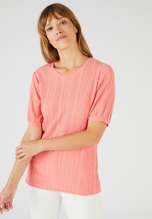 Donna con capelli castano chiaro che indossa una blusa corallo a maniche corte e con texture e pantaloni bianchi, in piedi davanti a uno sfondo semplice.