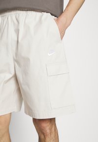 Světle béžové cargo šortky z lehkého materiálu, s elastickým pasem, bočními kapsami a malým bílým logem Nike na kapse.