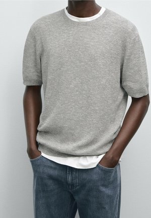 Mann trägt hellgrauen kurzärmeligen Strickpullover über weißem Hemd, Hände in den Taschen und blaue Jeans, vor einfachem Hintergrund.