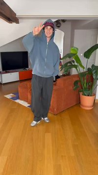 Lyseblå zip-hoodie, svarte baggy bukser og sølvfargede sko. Innendørs setting med en potteplante og en oransje sofa i bakgrunnen.
