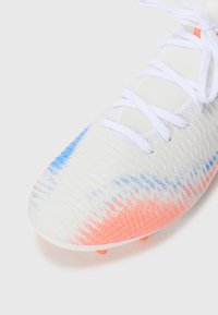 Botte blanche avec un design pixelisé orange et bleu sur le bout, surface texturée et lacets blancs sur un fond uni.