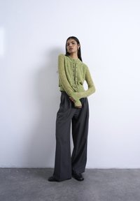 Γυναίκα με πράσινη πλεκτή ζακέτα με κουμπιά και φαρδιά σκουρόχρωμη γκρι παντελόνα στέκεται μπροστά σε ένα λευκό τοίχο με τα χέρια σταυρωμένα.