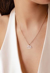 Collana con pendente a forma di loto in argento su una catena delicata, caratterizzata da un intricato design floreale. Indossata sopra una maglietta di tessuto bianco.