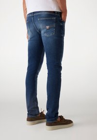 Blue denim jeans with a fitted design. Features two back pockets, a logo detail, and light fading on the thighs. Worn with brown shoes.