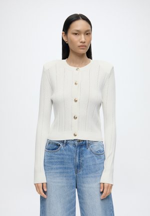 Femme portant un cardigan en maille blanc boutonné avec des épaules bouffantes et un jean taille haute bleu clair sur un fond blanc uni.