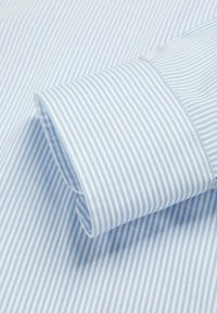 Puño de camisa a rayas verticales azul claro y blanco con costuras precisas sobre un fondo de tela de algodón.