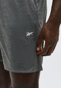 Άτομο με σκουρόχρωμο δέρμα που φοράει γκρι αθλητικό σορτς με λευκό λογότυπο Reebok, με το χέρι χαλαρό δίπλα στον μηρό, στέκεται μπροστά σε απλό φωτεινό φόντο.