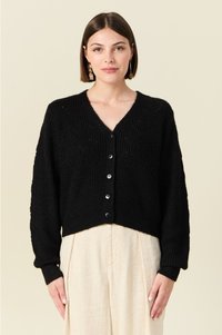 Cardigan noir en tricot avec un col en V, des manches longues et six boutons à l'avant. Présente un motif texturé et un ourlet court. À porter par-dessus un pantalon clair.
