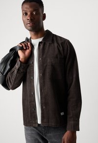 Brown corduroy shirt with vertical stripes, buttoned front, chest pocket, and a white undershirt. Model holds a black bag over one shoulder.