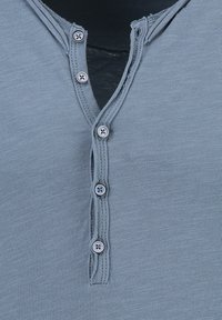 Grijze henley shirt met een v-hals, vijf knopen aan de voorkant en een textuurstof. Randen zijn lichtjes gewafeld.