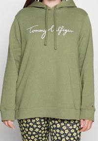 Oliven grønn hettegenser med hvit brodert logo, justerbar hette med snor og ribbestrikkede mansjetter. Kombinert med bukser med blomstermønster.