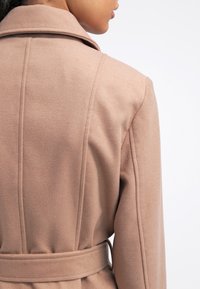 Kvinde iført en beige uldfrakke med krave og bælte, vist fra ryggen mod en ensfarvet hvid baggrund.