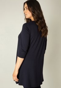 Marineblauwe tuniektop met driekwart mouwen, zachte stof, losse pasvorm en afgeronde zoom, ontworpen voor comfort en bewegingsvrijheid.