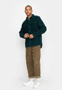 Donkergroen corduroy overhemdjack met twee borstzakken, knoopsluiting, gedragen over een lichtgroene top, gecombineerd met khaki corduroy broek en witte sneakers.