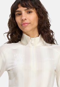 Dlhý rukáv s zipom na svetlo béžovej farbe a s vertikálnymi pruhmi, so vyšitým logom a golierom typu mock. Hladký, elastický materiál.