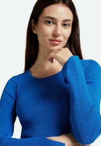 Top lungo blu a maniche lunghe con scollatura rotonda. Presenta un tessuto strutturato con sottili luccichii, forma aderente e maniche allungate.