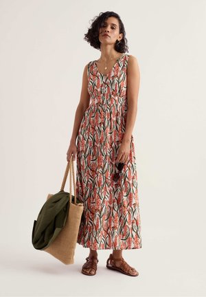 Žena v bezrukávových maxi šatách s kvetinovým vzorom, hnedých sandáloch, drží veľkú tkanú tašku so zelenou bundou vo vnútri a slnečné okuliare.