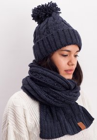 Knit dark gray beanie with pom-pom and matching scarf featuring ribbed texture and leather accent, worn over a light sweater.