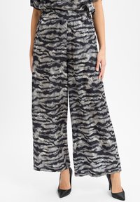 Vrouw in wijde broek met zwart/grijs abstract zebra patroon, gecombineerd met zwarte puntige hoge hakken.