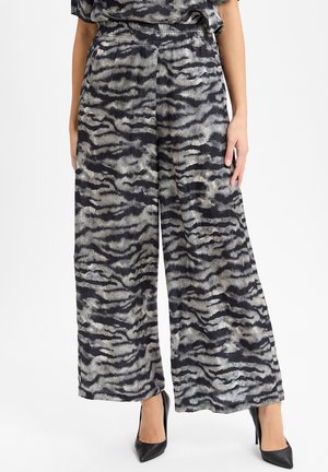 Vrouw in wijde broek met zwart/grijs abstract zebra patroon, gecombineerd met zwarte puntige hoge hakken.