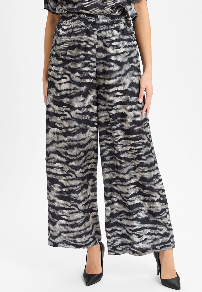 Vrouw in wijde broek met zwart/grijs abstract zebra patroon, gecombineerd met zwarte puntige hoge hakken.