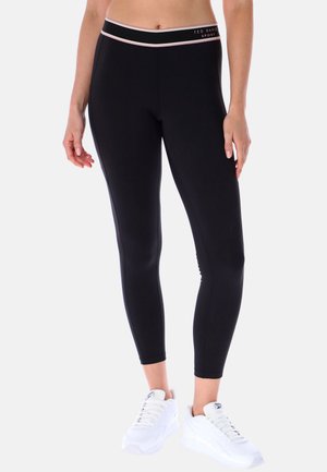 Femme portant des leggings noirs ajustés avec une fine ceinture rose portant la mention "Ted Baker Sport" et des baskets blanches.