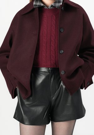 Femme portant un manteau bordeaux par-dessus un pull torsadé marron, une chemise à carreaux noirs et blancs, un short en cuir noir et des collants fins.