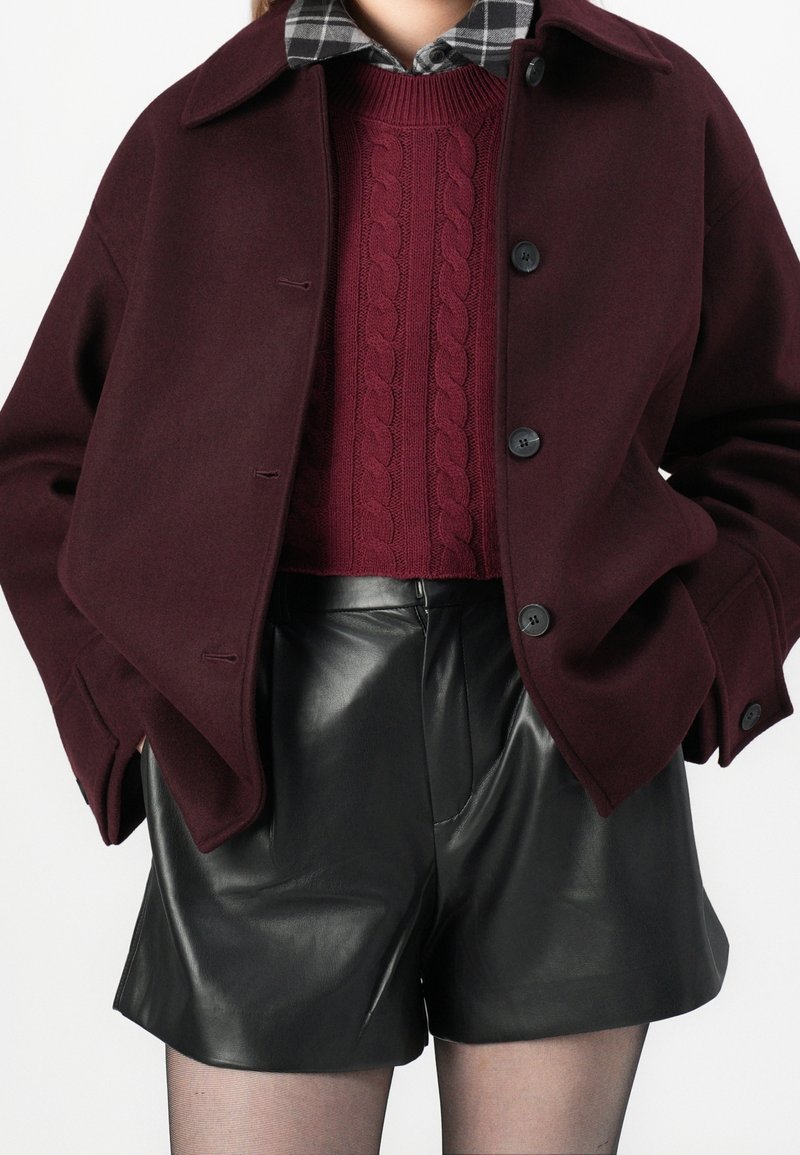 Femme portant un manteau bordeaux par-dessus un pull torsadé marron, une chemise à carreaux noirs et blancs, un short en cuir noir et des collants fins.