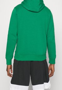 Personne portant un sweat à capuche vert et un short noir avec des bordures blanches, vu de dos sur un fond neutre.