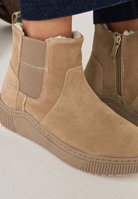 Bottines en suède beige avec des panneaux latéraux élastiques et une fermeture à glissière. Semelle en caoutchouc texturé avec des lignes horizontales pour l'adhérence et le soutien.