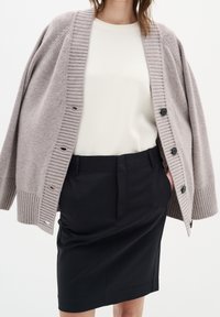 Cardigan beige clair avec des bords côtelés, agrémenté de boutons foncés ; superposé à un haut blanc et une jupe noire taille haute. Texture douce.