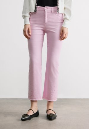 Femme portant un jean court rose orné de petites perles près des ourlets effilochés et des ballerines noires pointues avec des boucles dorées.