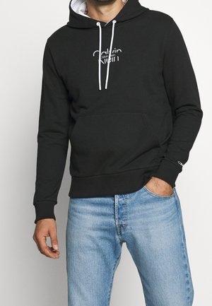 Man wearing black Calvin Klein hoodie with white drawstrings and light blue jeans, hand in pocket, cropped at shoulders and thighs.