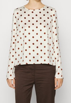 Witte blouse met lange mouwen met bruine stippen, gecombineerd met donkerbruine broek, getoond op een staande persoon tegen een effen achtergrond.